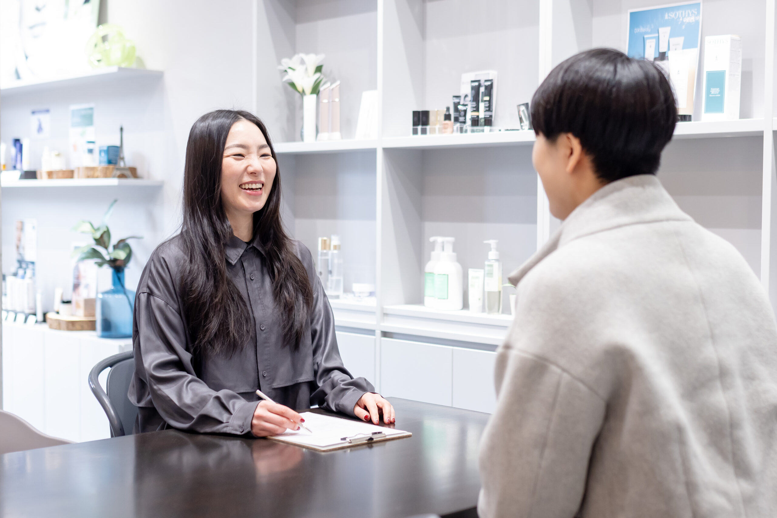 化粧品専門店の知識を活かして肌状態を見極める肌診断サポート - 三田市エステサロン ルナクレッシェンテのスタッフが化粧品棚を背景にお客様の肌質や悩みに合わせた施術とスキンケアを提案